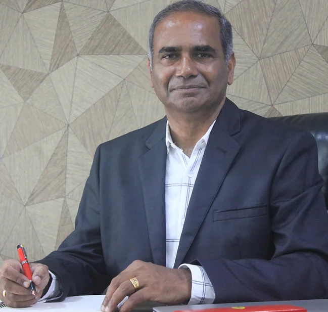 A man in a dark suit sits at a desk, signing with a red pen against a geometric beige wall background.