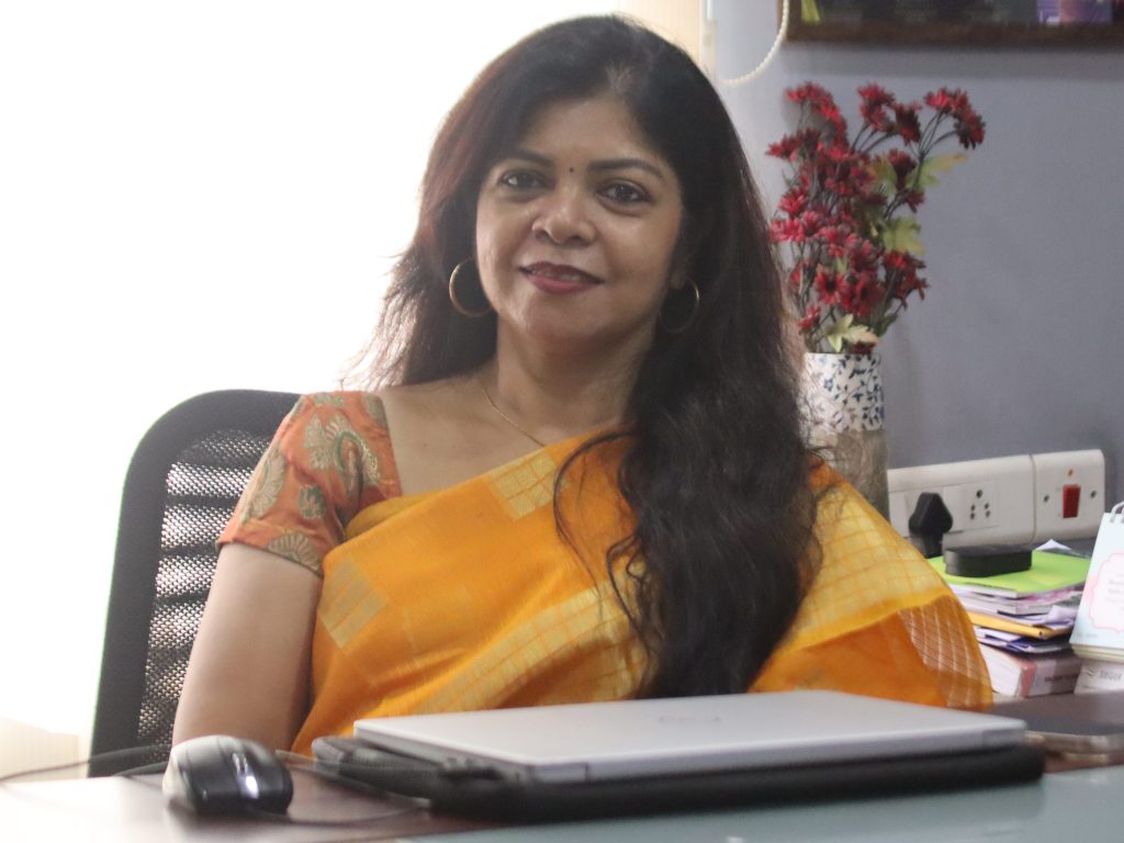 Woman seated at a desk in an office, wearing an orange sari, facing the camera with a slight smile, computer mouse and papers nearby.